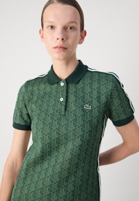 Polo verde cu un model geometric, cu mâneci scurte și guler. Include două butoane și dungi laterale contrastante.