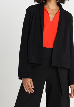 Femme portant un chemisier rouge col en V sous une veste noire à ouverture avant, assorti à un pantalon large noir taille haute.