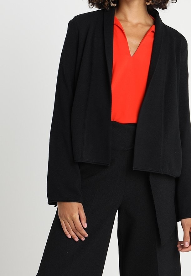 Femme portant un chemisier rouge col en V sous une veste noire à ouverture avant, assorti à un pantalon large noir taille haute.