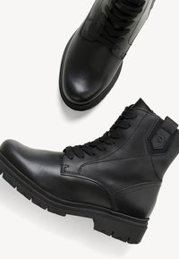 Botine negre din piele cu o finisare netedă, vârf rotund și șireturi în față. Prezintă părți texturate și o talpă din cauciuc negru rezistent.