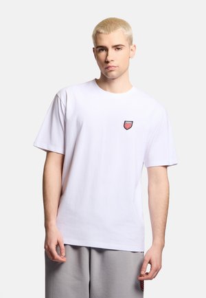 Młody mężczyzna z krótkimi blond włosami, ubrany w biały T-shirt z małym czerwono-czarnym logo w kształcie tarczy na piersi oraz jasnoszare spodnie.