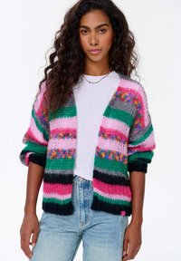 Vícebarevný pletený kardigan s pruhy v růžové, zelené, šedé a černé barvě, volného střihu s žebrovanými lemy, nošený přes bílou košili.