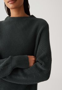 Donna che indossa un maglione a coste grigio scuro con maniche lunghe, con le braccia incrociate su uno sfondo chiaro e semplice.
