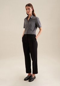 Camicia grigia a maniche corte con bottoni, pantaloni neri a vita alta e mocassini neri. Tessuto liscio, vestibilità sartoriale e tasca frontale visibile.