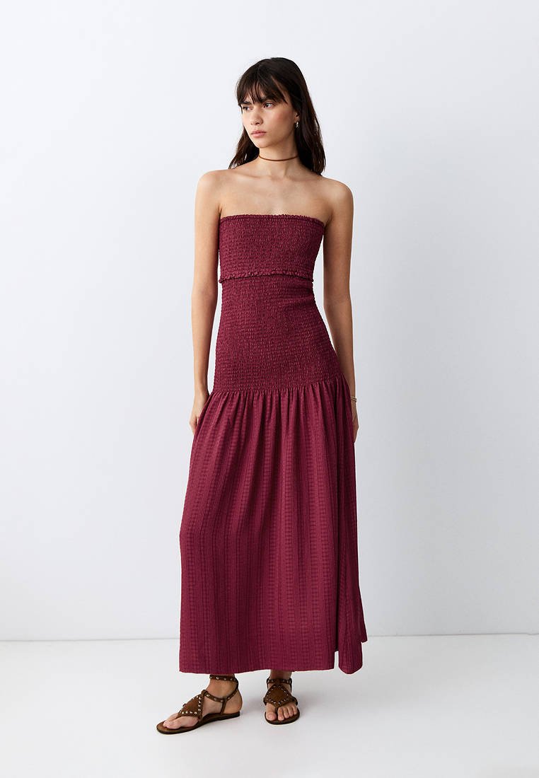 Femme portant une robe maxi bordeaux sans bretelles et texturée avec des sandales marron à clous, debout devant un fond blanc uni.