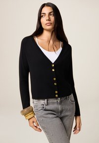 Schwarze Strickjacke mit V-Ausschnitt und goldenen Knöpfen, kombiniert mit grauen Jeans mit Verzierungen. Glatter Stoff mit taillierter Passform.