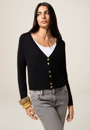 Zwarte cardigan met een V-hals en gouden knopen, gecombineerd met grijze jeans met versieringen. Gladde stof met een getailleerd ontwerp.
