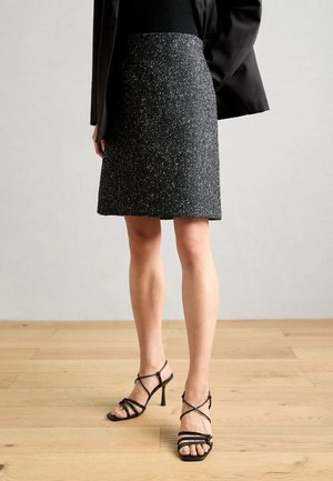 Mujer con falda de patrón en blanco y negro, blazer negro y sandalias negras de tacón alto con tiras, de pie sobre suelo de madera.