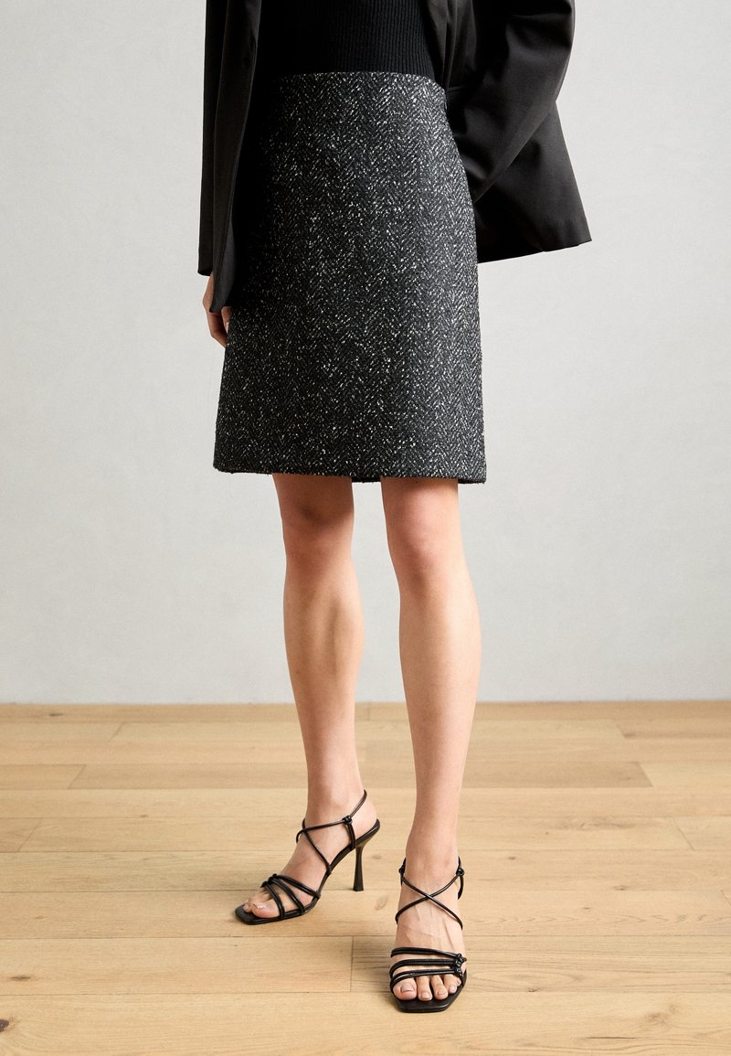 Femme portant une jupe à motifs noir et blanc, un blazer noir et des sandales à talons hauts noires à brides, debout sur un sol en bois.
