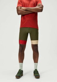 Heren fietsbroeken in groen met rode en beige accenten, vervaardigd uit rekbaar materiaal. De set omvat een rood shirt en groene sokken met een patroon.