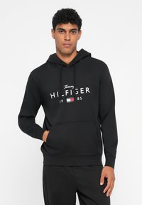 Sweatshirt preto feito de um tecido suave, com um bolso frontal, capuz com cordão e logótipo branco "Tommy Hilfiger" com um pequeno detalhe de bandeira.