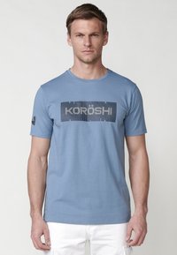 Svijetloplava pamučna majica kratkih rukava, s grafičkim dizajnom "KORÖSH" u bijeloj i tamnoplavoj boji. Okrugli izrez.