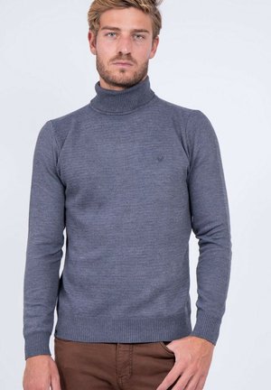Homme blond portant un pull à col roulé gris moulant et un pantalon marron, debout devant un fond clair uni.