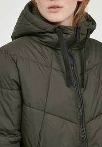 Detailní záběr osoby ve vojensko zelené prošívané bundě s vysokým límcem, zapínáním na zip a černými šňůrkami na kapuci.