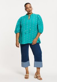 Tegen een turquoise blouse met opgeblazen mouwen, een geknoopte halslijn en bloemenborduursels. Gepaard met cropped denim broekjes met een contrasterende zoom.