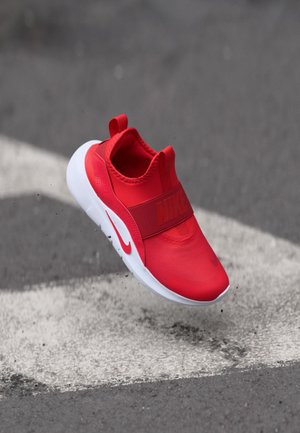 Rote Nike Slip-On-Sneaker haben ein strukturiertes Obermaterial, einen breiten Riemen mit Logo und eine weiße Gummisohle. Sie werden in der Luft über einem Gehweg gezeigt.