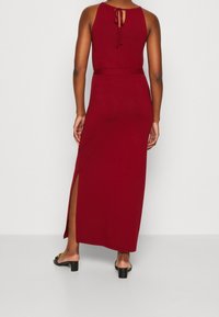 Robe maxi en tissu rouge foncé avec une taille ajustée, un détail de lien dans le dos et une fente sur le côté. Présente un design épuré avec une texture lisse.