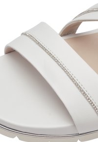 Vita sandaler har breda, släta korsremmar med en metallisk kant av små rhinestones längs mitten och en dämpad fotbädd.