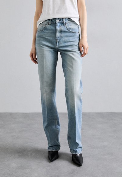 Jean droit en denim bleu clair avec un subtil design délavé, stylé avec cinq poches et une fermeture à bouton, associé à des chaussures noires à bout pointu.
