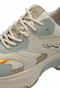 Beige und hellgrauer Sneaker aus Mesh- und Leder-Materialien, mit metallischen Akzenten, einem strukturierten goldenen Bereich und beigen Schnürsenkeln.