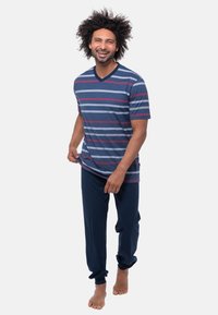 Heren T-shirt met V-hals in blauw met roze en witte horizontale strepen, gecombineerd met donkerblauwe broeken. Zachte stof, relaxte pasvorm. Blootsvoets.