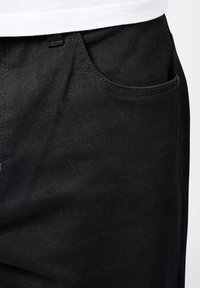 Pantalones de mezclilla negros con una textura suave, que cuentan con un bolsillo frontal y un corte clásico. La cinturilla incluye un pequeño lazo para un detalle adicional.
