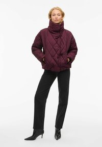 Quiltana jakna boje burgundca s visokim ovratnikom, detaljem za stezanje i dva prednja džepa, uparen s crnim trapericama ravnog kroja i čizmama do gležnja.