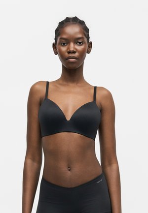 Jonge zwarte vrouw met gevlochten haar, een zwarte beha en bijpassend ondergoed tegen een effen witte achtergrond.