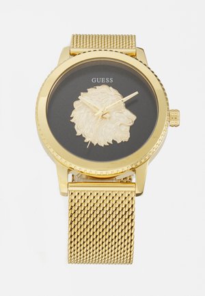 Montre dorée avec un bracelet en maille, cadran noir présentant un détail en forme de tête de lion, et aiguilles des heures et des minutes dorées. Marque affichée en haut.