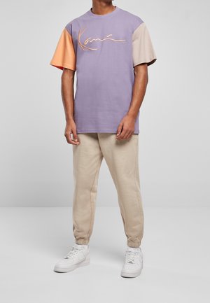 Lilla kortærmet t-shirt med orange og beige accenter, prydet med et kursiv logo. Matchende beige joggers og hvide sneakers.
