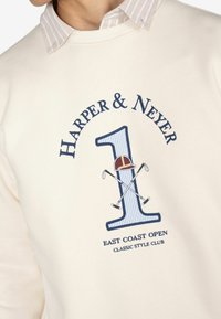 Sudadera color crema con un gráfico azul del "1", texto "HARPER & NEYER" y palos de golf cruzados, con un cuello de rayas y detalles bordados.