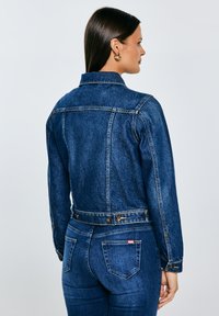 Moteris su ilgais tamsiais plaukais, dėvinti auksinius žiedinius auskarus, mėlyną džinsinę striukę ir mėlynas džinsines kelnes, atsukta nugarą priešais paprastą foną.