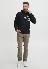 Felpa nera con testo bianco, tasca frontale e cappuccio con cordino, abbinata a pantaloni khaki e sneakers bianche, in piedi su uno sfondo neutro.