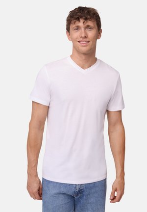 Nuori mies kiharatukkaisena, pukeutunut yksinkertaiseen valkoiseen V-kaulaiseen T-paitaan ja sinisiin farkkuihin, seisoo vaaleaa taustaa vasten ja hymyilee.