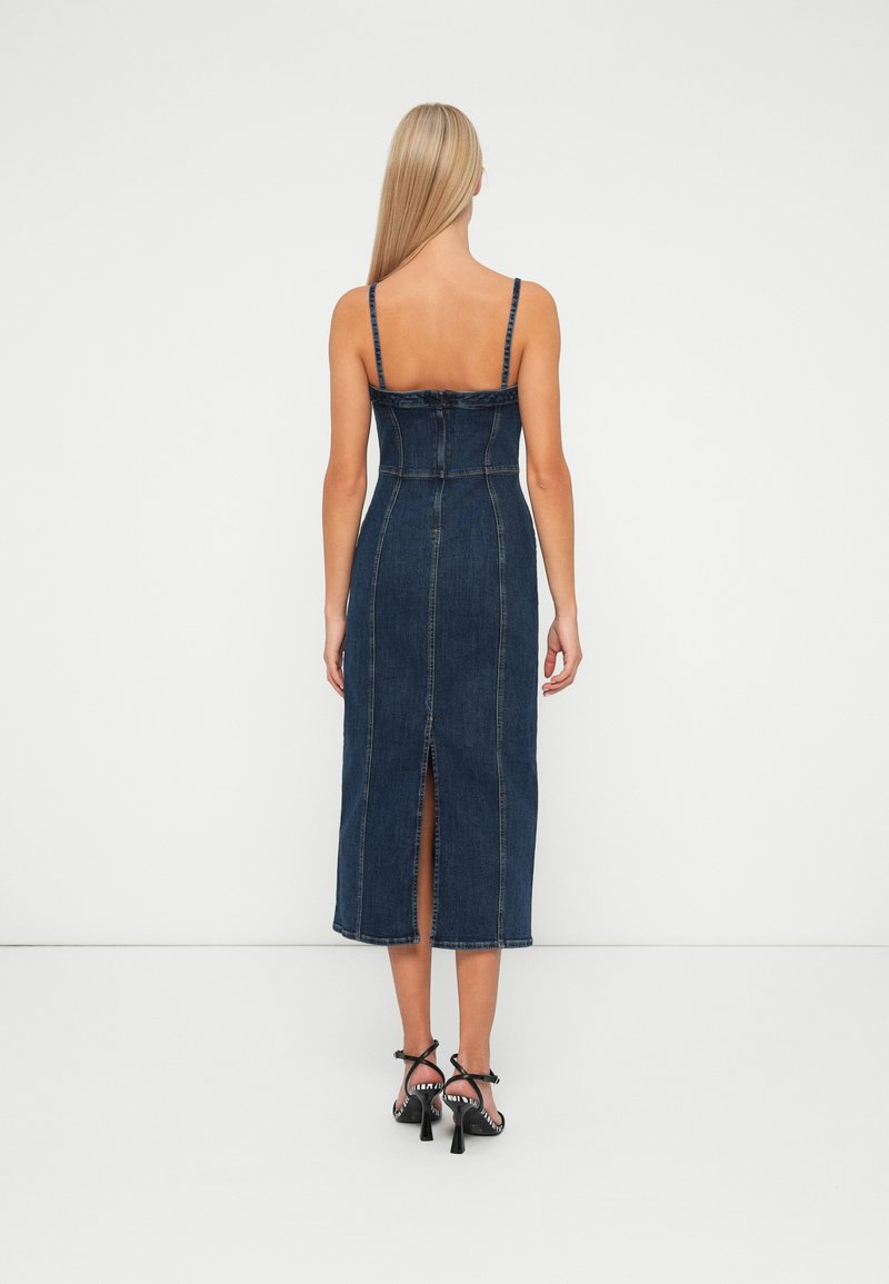 Vrouw met lang blond haar in een donkerblauwe denim midi-jurk met spaghettibandjes en een split aan de achterkant, gecombineerd met zwarte sandaaltjes met hakken.