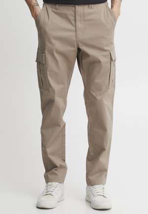 Man som bär beige cargobyxor och vita sneakers, står med händerna i fickorna mot en neutral bakgrund.