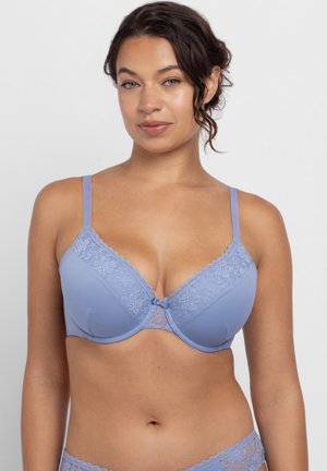 Γυναίκα που φοράει ανοιχτό μπλε δαντελένιο σουτιέν και ταιριαστό εσώρουχο, κοιτάζει μπροστά με ουδέτερη έκφραση σε απλό φόντο.