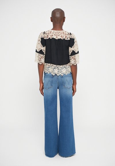 Top in pizzo nero e beige con maniche larghe, caratterizzato da motivi intricati. Abbinato a jeans in denim blu a zampa d'elefante con dettagli di cuciture in vista.