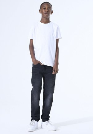 Witte korte mouwen t-shirt, donkerblauwe jeans en witte sneakers. Model staat met één hand in de zak tegen een effen achtergrond.