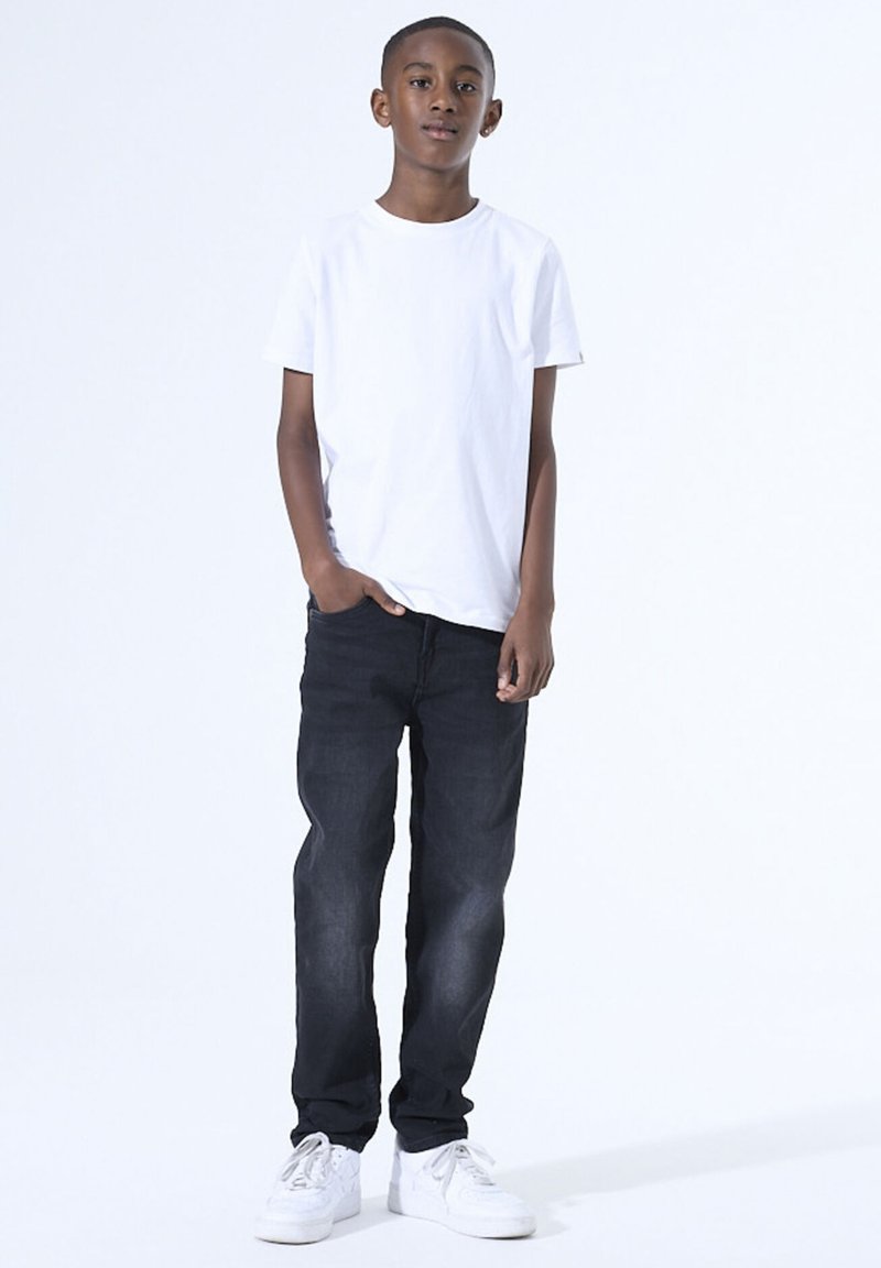 Witte korte mouwen t-shirt, donkerblauwe jeans en witte sneakers. Model staat met één hand in de zak tegen een effen achtergrond.