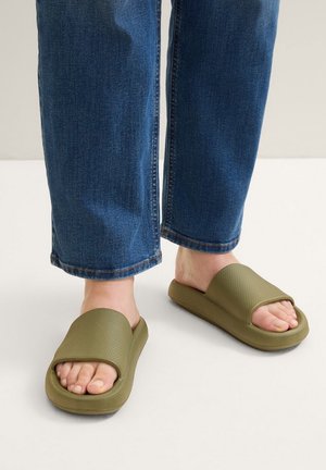 Des pieds portant des sandales à glissière texturées olive avec de larges sangles, associées à un jean en denim bleu raccourci au-dessus des chevilles.