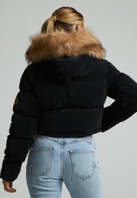 Zwarte pufferjack met een bontkap, met een cropped ontwerp en zichtbare stiksels. Draagt over lichtblauwe spijkerbroek.