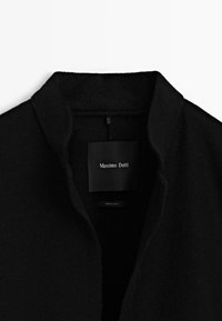 Collo di un cappotto di lana nero con l'etichetta del marchio "Massimo Dutti" cucita all'interno, fotografato su uno sfondo bianco.