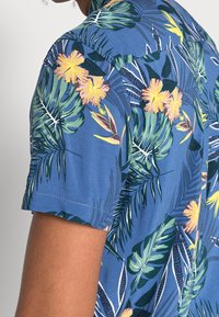 Camicetta a maniche corte in tessuto blu con una stampa tropicale di foglie verdi e fiori rosa-gialli, con dettagli di cucitura.