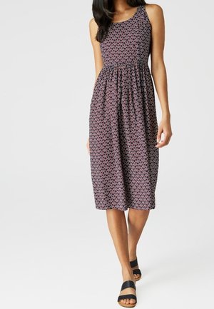 Femme portant une robe sans manches mi-longue avec un motif géométrique violet et noir et des sandales noires ouvertes, marchant vers l'avant sur un fond uni.