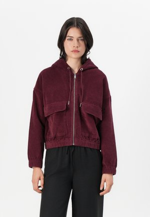 Veste à capuche en velours côtelé bordeaux avec deux poches avant, fermeture éclair et taille élastique. Texture lisse avec une coupe oversize.
