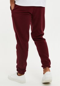 Bordeaux fleece joggingbroek met elastische boorden, zijzakken en een comfortabele pasvorm, gecombineerd met witte sneakers. De nadruk ligt op comfort en functionaliteit.
