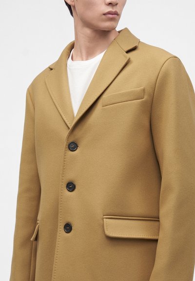 Homme portant un manteau beige boutonné par-dessus une chemise blanche, regardant légèrement sur le côté, sur un fond clair uni.