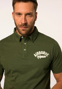 Zelené polo tričko s límečkem, na kterém je bíle vyšívaný text "AIRBORNE Heroes", s knoflíkovým zapínáním a příjemnou mírně měkkou texturou.