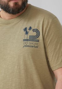 Olijfgroene t-shirt met korte mouwen van zachte stof, met een marineblauw grafisch ontwerp van palmbomen en de tekst "OFF SHORE pleasures."